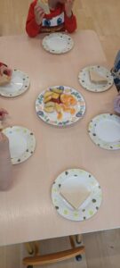 Na zdjęciu widać stół, przy którym siedzi kilkoro dzieci. Na stole ustawiono sześć papierowych talerzyków — na każdym z nich leży trójkątne kawałki ciasta Pischinger z kajmakiem. Na środku stołu znajduje się większy talerz z pokrojonymi owocami, m.in. kawałkami mandarynek i bananów. 