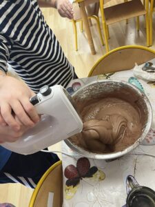 Na zdjęciu widać dziecko w pasiastym, czarno-białym swetrze, które trzyma mikser ręczny i miesza czekoladowe ciasto w dużej metalowej misce. Miska stoi na stole przykrytym ceratą z motywem motyli. W tle widać żółte krzesła i fragment sali, prawdopodobnie przedszkolnej lub szkolnej. Masa w misce jest gładka i kremowa, wygląda jak przygotowywane ciasto na babeczki lub ciasto czekoladowe. Scena przedstawia moment wspólnego pieczenia lub zajęć kulinarnych.