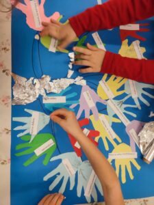 Na zdjęciu widać pracę plastyczną tworzoną przez dzieci. Na dużym niebieskim kartonie przyklejone są kolorowe wycinanki w kształcie dłoni z zapisanymi na nich miłymi słowami i komplementami, takimi jak „Dzień dobry”, „Jesteś super”, „Masz w sobie SUPERmoc!”, „Miło cię widzieć”. Dzieci doklejają do pracy kawałki srebrnej folii, tworząc dodatkowe elementy dekoracyjne. Całość wygląda na wspólny projekt związany z życzliwością lub pozytywnymi komunikatami.