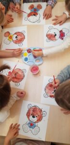 Na zdjęciu widać grupę dzieci siedzących przy stole i malujących obrazki przedstawiające misia. Każde dziecko ma przed sobą kartkę z tym samym rysunkiem – uśmiechniętym misiem z dużą głową, serduszkiem na brzuszku i kokardką lub guzikiem.

Dzieci używają farb akwarelowych w odcieniach brązu, czerwieni i żółci, a na środku stołu stoi paleta z farbami oraz kubeczek z wodą do płukania pędzli. Każde dziecko skupia się na swojej pracy, starannie kolorując misia.

Scena ma ciepły, radosny charakter i wygląda na zajęcia plastyczne w przedszkolu, prawdopodobnie z okazji Dnia Pluszowego Misia.