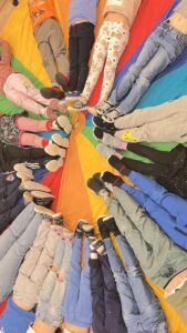 Na zdjęciu widać grupę dzieci leżących w kręgu na kolorowej chuście animacyjnej w barwach tęczy — czerwonej, pomarańczowej, żółtej, zielonej, niebieskiej i fioletowej. Dzieci ułożyły się tak, że ich nogi skierowane są do środka, tworząc wzór przypominający promienie słońca. Mają na sobie różnorodne ubrania — jeansy, legginsy, dresy i kolorowe skarpetki. Scena wygląda radośnie i dynamicznie, jak element zabawy grupowej w przedszkolu lub podczas zajęć integracyjnych.