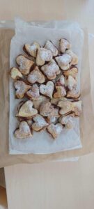 Na zdjęciu widać blachę lub tacę wyłożoną papierem do pieczenia, na której ułożone są ciasteczka w kształcie serduszek. Ciasteczka są złociste, lekko przyrumienione na brzegach i posypane cukrem pudrem. Wyglądają na kruche lub francuskie, a między warstwami widać nadzienie — prawdopodobnie dżem lub marmoladę. Całość prezentuje się apetycznie i domowo, jak wypieki przygotowane na specjalną okazję, np. dzień życzliwości.