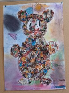 Na zdjęciu widać dużą pracę plastyczną przedstawiającą misia wykonanego techniką kolażu. Sylwetka misia została wypełniona wydartymi kawałkami kolorowych gazet i papieru, które tworzą ciekawą, fakturowaną powierzchnię. Fragmenty papieru mają różne barwy – przeważają odcienie brązu, pomarańczu, czerwieni i niebieskiego, co nadaje postaci żywy, patchworkowy wygląd.

Tło pracy jest pokolorowane pastelami lub kredkami w delikatnych odcieniach fioletu, zieleni, żółci i błękitu. W prawym górnym rogu narysowane jest słońce, a po lewej stronie fioletowe serduszko. W dolnej części widać dziecięce rysunki – linie i kształty dodane spontanicznie, co podkreśla twórczy charakter pracy.

Całość wygląda radośnie i ekspresyjnie, a misio sprawia wrażenie przyjaznego i pełnego energii. To przykład kreatywnej pracy grupowej lub indywidualnej wykonanej przez dziecko w przedszkolu