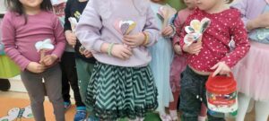 Na zdjęciu widać grupę kilku dzieci stojących obok siebie w sali przedszkolnej. Dzieci trzymają w rękach papierowe kwiatki wykonane z kolorowych serduszek naklejonych na patyczki. Jedno z dzieci trzyma czerwone pudełko z rączką. Atmosfera wygląda na radosną, jak podczas zajęć plastycznych lub święta w przedszkolu.