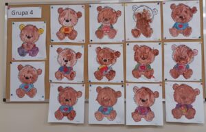 Na zdjęciu widać tablicę korkową z pracami plastycznymi dzieci z grupy 4. Na tablicy przypięto piętnaście rysunków misiów, każdy wykonany przez inne dziecko.

Wszystkie prace przedstawiają tego samego misia – siedzącego, uśmiechniętego, z dużą głową, serduszkiem na brzuszku i kokardką pod szyją. Dzieci pomalowały misie farbami, głównie w odcieniach brązu, różu, żółci i niebieskiego. Każdy miś wygląda nieco inaczej – różnią się intensywnością kolorów, sposobem malowania i detalami, co nadaje całości uroku i indywidualnego charakteru.

Pod każdym rysunkiem widnieje imię dziecka, które go wykonało. Prace są starannie przypięte kolorowymi pinezkami, tworząc radosną, kolorową galerię przedszkolnych dzieł z okazji Dnia Pluszowego Misia.