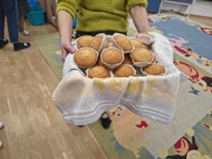 Na zdjęciu widać osobę trzymającą koszyk lub miskę przykrytą ściereczką, w której znajduje się kilkanaście upieczonych babeczek w papierowych foremkach. Babeczki są złociste i wyglądają na świeżo upieczone. 