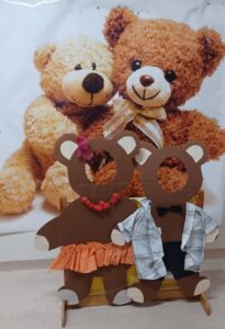 Na zdjęciu widać dekorację z okazji Dnia Pluszowego Misia. Na pierwszym planie stoją dwie duże, kartonowe sylwetki misiów z wyciętymi otworami na twarze, w które można włożyć głowę i zrobić pamiątkowe zdjęcie.

Lewy miś jest „dziewczynką” – ma różową kokardkę na uchu, naszyjnik z czerwonych koralików i pomarańczową spódniczkę z falbanką. Prawy miś to „chłopiec” – ubrany w koszulę w kratkę, czarne spodenki i muszkę.

W tle znajduje się duży plakat przedstawiający dwa pluszowe misie – jeden jasnobeżowy, drugi brązowy – siedzące obok siebie i uśmiechnięte. Całość tworzy ciepłą, przyjazną i radosną scenę, idealną do przedszkolnej sesji zdjęciowej lub zabawy tematycznej.