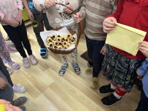 Na zdjęciu widać grupę dzieci stojących w kółku. Jedno z nich trzyma wiklinowy koszyk wypełniony małymi ciasteczkami z owocową konfiturą, a inne dziecko trzyma żółtą kopertę. Wszyscy mają kolorowe ubrania i kapcie, co sugeruje, że znajdują się w przedszkolu. Scena wygląda na radosną i związaną z jakąś wspólną aktywnością lub poczęstunkiem.