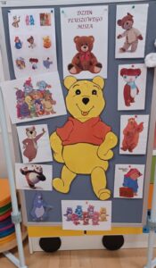 Na zdjęciu widać tablicę dekoracyjną przygotowaną z okazji Dnia Pluszowego Misia. W centralnej części znajduje się duży, kolorowy rysunek Kubusia Puchatka w czerwonej koszulce, który uśmiecha się i trzyma łapki uniesione w górę.

Wokół niego rozmieszczono ilustracje różnych znanych misiów z bajek i filmów, m.in. misia Paddingtona z walizeczką, misia z niebieskim futerkiem, misia z kreskówki o przyjaciołach z lasu, misia z grupy Care Bears oraz pandę. Na górze tablicy widnieje napis „DZIEŃ PLUSZOWEGO MISIA”, a obok niego zdjęcie klasycznego pluszowego misia w brązowym kolorze.