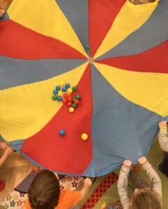 Na zdjęciu widać kolorową chustę animacyjną w barwach czerwonej, żółtej i niebieskiej, trzymaną przez kilkoro dzieci. Na chuście leży kilka małych, kolorowych piłeczek — głównie niebieskich, czerwonych, zielonych i żółtych. Dzieci stoją wokół chusty, każde trzyma jej brzeg obiema rękami. Scena wygląda na zabawę grupową w przedszkolu lub sali zabaw, a podłogę zdobi kolorowy dywan. Atmosfera jest radosna i pełna ruchu.