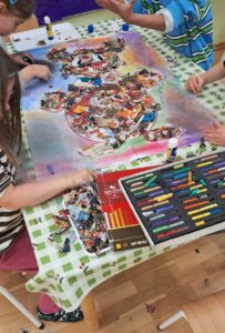 Na zdjęciu widać grupę dzieci siedzących przy stole i wykonujących wspólną pracę plastyczną. Na stole leży duży arkusz papieru, na którym powstaje kolorowa kompozycja przypominająca sylwetkę misia. Dzieci tworzą ją z wydartych kawałków kolorowych gazet i papieru, które przyklejają klejem w sztyfcie.

Tło pracy jest pokolorowane pastelami w różnych odcieniach — fioletowym, niebieskim, różowym i żółtym. Obok arkusza leży zestaw pastelowych kredek w pudełku oraz pudełko z resztkami papierowych skrawków.

Stół przykryty jest ceratą w zieloną kratkę, a dzieci są skupione i zaangażowane w swoją twórczość. Cała scena ma radosny, kreatywny charakter i wygląda na zajęcia plastyczne w przedszkolu lub szkole.