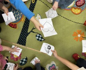 Na zdjęciu widać grupę dzieci siedzących lub klęczących w kręgu na zielonym dywanie z kolorowymi wzorami kwiatów i liści. Każde dziecko trzyma w rękach kartkę papieru z prostym rysunkiem — widać m.in. serca, postacie i inne kształty narysowane czarnym markerem. Dzieci mają kolorowe ubrania, a ich dłonie są wyciągnięte w kierunku środka kręgu, jakby pokazywały swoje prace. Scena wygląda na zajęcia plastyczne lub zabawę w przedszkolu.