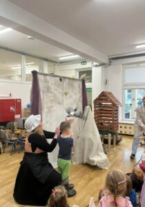 Na zdjęciu widać scenę z przedstawienia lub zajęć edukacyjnych w przedszkolu. Na pierwszym planie siedzi grupa dzieci, które z zainteresowaniem obserwują występ.

W centrum znajduje się kobieta ubrana w czarną suknię, biały czepek i fartuch, klęcząca obok małego chłopca w zielonych spodniach i niebieskiej koszulce. Kobieta trzyma w dłoniach figurkę gołębia, którą unosi w stronę zawieszonego tła z jasnej tkaniny.

Za nimi stoi drewniana konstrukcja przypominająca domek lub karmnik dla ptaków, a obok widać drugą osobę w jasnym stroju, częściowo zasłoniętą. Scena rozgrywa się w jasnej sali z dużymi oknami i dziecięcymi meblami.

Całość wygląda na fragment interaktywnego przedstawienia o ptakach, w którym dzieci biorą czynny udział, ucząc się poprzez zabawę.