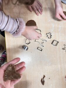 Na zdjęciu widać dzieci przy stole, które wycinają kształty z ciasta przy pomocy metalowych foremek. Na blacie rozsypana jest mąka, a widać też kawałki ciemnego ciasta, prawdopodobnie piernikowego.

Dzieci używają foremek w różnych kształtach – m.in. serca, kwadratu, kwiatka i zwierzątka. Jedno z dzieci trzyma w dłoni wycięte serce z ciasta, a inne sięga po foremkę w kształcie renifera.

Scena przedstawia zajęcia kulinarne lub świąteczne pieczenie ciasteczek, w których dzieci biorą aktywny udział, ucząc się i bawiąc jednocześnie.