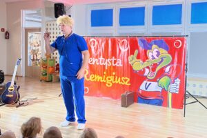 Na zdjęciu widać mężczyznę występującego przed grupą dzieci w sali przedszkolnej. Ma na sobie niebieski strój przypominający ubranie medyczne oraz jasną perukę. Stoi na scenie obok czerwonego baneru z napisem „Dentysta Remigiusz” i dużą, wesołą ilustracją zielonego krokodyla w czapce i koszulce. Obok stoi gitara na statywie i mały wzmacniacz. Dzieci siedzą na podłodze i patrzą na występ. Atmosfera jest kolorowa i radosna.