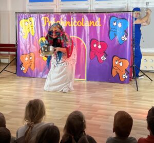 Na zdjęciu widać scenkę teatralną wystawianą dla dzieci. Na pierwszym planie siedzi grupa maluchów, które z zainteresowaniem oglądają przedstawienie. Na scenie znajduje się osoba przebrana w kolorowy strój — ma na sobie dużą chustę, ogromne okulary i trzyma metalową miskę. W tle widać kolorową dekorację z napisem „Próchnicoland” oraz rysunkami zębów o różnych wyrazach twarzy — niektóre wyglądają na smutne lub przestraszone. Po prawej stronie stoi inna osoba ubrana na niebiesko, prawdopodobnie odgrywająca rolę dentysty lub bohatera walczącego z próchnicą. Całość wygląda jak edukacyjne przedstawienie o higienie jamy ustnej dla dzieci.