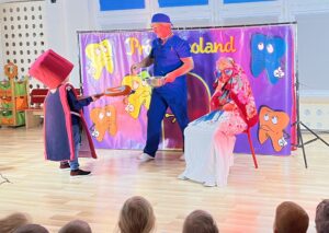 Na zdjęciu widać scenę przedstawienia teatralnego dla dzieci. Na środku stoi mężczyzna ubrany w niebieski strój przypominający fartuch dentysty i czapkę chirurgiczną. W rękach trzyma metalową miskę i łyżkę. Po jego lewej stronie stoi dziecko przebrane w kostium z dużym czerwonym cylindrem na głowie i peleryną — wygląda jak postać z bajki lub symbol próchnicy. Po prawej stronie siedzi osoba w kolorowej peruce, dużych okularach i chuście, przykryta białym materiałem, odgrywająca zapewne pacjenta lub zabawną postać.