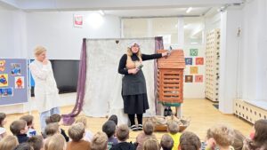 Na zdjęciu widać scenę przedstawienia lub zajęć edukacyjnych w przedszkolu. Na pierwszym planie siedzi grupa dzieci zwróconych w stronę dwóch kobiet stojących przed nimi.
Po prawej stronie stoi kobieta ubrana w czarną sukienkę, fartuch i biały czepek, trzymająca w dłoniach dwa gołębie – jednego przy piersi, a drugiego uniesionego w górę. Obok niej znajduje się drewniana konstrukcja przypominająca domek lub karmnik dla ptaków, w której widać gniazdo z gałązek i gołębie.