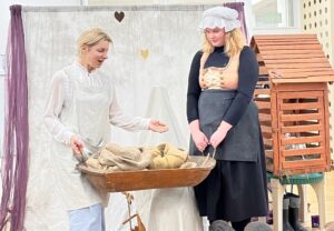 Na zdjęciu widać dwie kobiety biorące udział w przedstawieniu lub zajęciach edukacyjnych, prawdopodobnie w przedszkolu lub szkole.

Po lewej stronie stoi kobieta ubrana w jasny strój – białą bluzkę i fartuch, z niebieskimi spodniami. W dłoniach trzyma drewnianą miskę lub koryto wypełnione workami z jutowego materiału, które wyglądają jak woreczki zboża lub mąki. Kobieta gestykuluje, jakby coś tłumaczyła lub opowiadała.

Po prawej stronie stoi druga kobieta w czarnej sukni, fartuchu i białym czepku, z gorsetem w odcieniach beżu i złota. Obserwuje pierwszą z uśmiechem, trzymając ręce splecione przed sobą.

W tle znajduje się jasna tkanina zawieszona jak kurtyna z ozdobnymi serduszkami oraz drewniana konstrukcja przypominająca domek lub karmnik dla ptaków. Na dole widać głowy dzieci, które oglądają scenę.

Całość sprawia wrażenie teatralnego przedstawienia o dawnych czasach lub pracy na wsi, prowadzonego w sposób edukacyjny i angażujący.