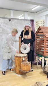 Na zdjęciu widać dwie kobiety biorące udział w przedstawieniu lub pokazie edukacyjnym, prawdopodobnie w przedszkolu. Obie ubrane są w stroje stylizowane na dawne — jedna ma na sobie czarną suknię, gorset w kolorze beżowo-złotym i biały czepek, a druga jasny fartuch i błękitne spodnie.

Kobiety stoją przy drewnianym urządzeniu przypominającym żarna lub młynek do mielenia zboża. Jedna z nich trzyma okrągły kamień młyński, a druga wskazuje na wnętrze naczynia, w którym widać rozsypaną mąkę. Obok znajduje się drewniane wiaderko oraz duża konstrukcja przypominająca domek lub karmnik dla ptaków.

W tle widać jasną tkaninę zawieszoną jak kurtynę oraz fragment sali z oknami i fioletową zasłoną. Na dole kadru widać głowy dzieci, które obserwują pokaz. Scena przedstawia demonstrowanie tradycyjnego sposobu mielenia zboża, w formie edukacyjnej i teatralnej.