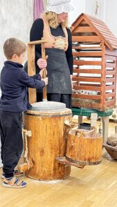 Na zdjęciu widać kobietę i chłopca uczestniczących w pokazie lub zajęciach edukacyjnych o dawnych rzemiosłach.

Kobieta ubrana jest w czarną suknię, fartuch, beżowo-złoty gorset i biały czepek. Obok niej stoi drewniane urządzenie przypominające żarna lub młynek do mielenia zboża. Chłopiec, ubrany w granatową bluzę, czarne spodnie i sandały, trzyma pionowy uchwyt i pomaga w kręceniu mechanizmem.

W tle widać drewnianą konstrukcję przypominającą domek lub karmnik dla ptaków, ustawioną na stole, a obok kilka naczyń i rekwizytów z drewna. Cała scena odbywa się w jasnej sali z drewnianą podłogą i wygląda na pokaz edukacyjny o tradycyjnym mieleniu zboża lub pracy na wsi.