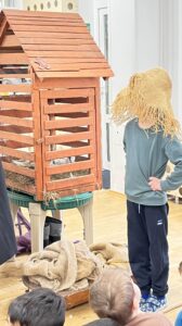 Na zdjęciu widać dziecko biorące udział w przedstawieniu lub zabawie teatralnej w sali przedszkolnej. Dziecko stoi obok drewnianej konstrukcji przypominającej domek lub karmnik dla ptaków, ustawionej na stole. W środku widać siano i elementy dekoracyjne.

Dziecko ma na sobie zieloną bluzę, granatowe spodnie i niebieskie buty, a na głowie duży kapelusz ze słomy, który zasłania mu twarz – wygląda jak element kostiumu, prawdopodobnie przedstawiający stracha na wróble.

Na podłodze leżą worki jutowe i kawałki materiału, a przed sceną siedzi grupa dzieci obserwujących występ. Całość ma charakter zabawnej, edukacyjnej inscenizacji o tematyce wiejskiej lub przyrodniczej.