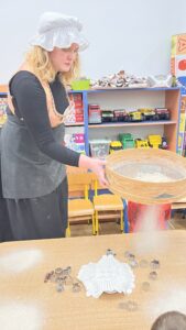 Na zdjęciu widać kobietę ubraną w czarną suknię, fartuch, beżowo-złoty gorset i biały czepek, stylizowaną na postać z dawnych czasów. Stoi przy stole i przesiewa mąkę przez duże, okrągłe sito, tworząc delikatną chmurkę białego pyłu.

Na stole przed nią leży biała, szydełkowa serwetka oraz kilka metalowych foremek do ciastek w różnych kształtach. W tle widać regał z zabawkami i modelami samochodów, co sugeruje, że scena odbywa się w sali przedszkolnej lub szkolnej.

Całość przedstawia pokaz edukacyjny o dawnym pieczeniu lub przygotowywaniu mąki, prowadzony w sposób ciekawy i angażujący dzieci.