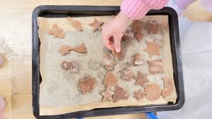 Na zdjęciu widać blachę do pieczenia wyłożoną papierem, na której ułożone są surowe ciasteczka z ciemnego ciasta, prawdopodobnie piernikowego. Ciasteczka mają różne kształty – gwiazdek, serc, kwiatków, choinki, ryby, księżyca i kółka z napisem.

W górnej części zdjęcia widać dziecięcą dłoń w różowym swetrze, która sięga po jedno z ciastek, jakby poprawiała jego ułożenie. Tło stanowi drewniany blat stołu lub podłogi, a obok widoczne są fragmenty ubrań dzieci.

Scena przedstawia przygotowania do pieczenia ciasteczek, prawdopodobnie podczas świątecznych zajęć kulinarnych dla dzieci.