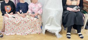 Na zdjęciu widać cztery osoby siedzące obok siebie na krzesłach, przykryte dużymi kocami i tkaninami w kwiatowe wzory. Trzy z nich to dzieci, a czwarta osoba dorosła siedzi po prawej stronie.
Każde dziecko trzyma w dłoniach figurkę gołębia, a dorosła osoba również ma jednego ptaka na kolanach. Dzieci ubrane są w kolorowe bluzy – granatową, różową i czarną – natomiast dorosła osoba ma na sobie czarną spódnicę, czarne rajstopy i gorset w odcieniach beżu i złota.
Podłoga jest drewniana, a tło stanowi jasna tkanina zawieszona za uczestnikami. Scena wygląda na fragment zajęć edukacyjnych lub przedstawienia o ptakach, w którym dzieci biorą aktywny udział, trzymając rekwizyty.