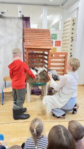 Na zdjęciu widać scenkę przedstawioną w przedszkolu. Na pierwszym planie siedzi kilkoro dzieci, które obserwują występ.

W centrum znajduje się drewniana konstrukcja przypominająca domek lub karmnik dla bocianów, ustawiona na stole. W środku widać gniazdo z gałązek i sztuczne bociany. Obok konstrukcji klęczy kobieta w jasnym fartuchu i niebieskich spodniach, trzymająca jednego z bocianów-pacynek. Przed nią stoi chłopiec w czerwonej bluzie i kaloszach, który również uczestniczy w przedstawieniu, wskazując coś w gnieździe.

W tle widać dekoracje sali – kolorowe prace dzieci na ścianie i zasłonę teatralną. Scena wygląda na fragment przedstawienia edukacyjnego lub teatralnego o bocianach i ich gnieździe, prowadzonego w sposób interaktywny z udziałem dzieci.