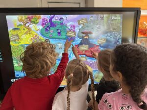 Na zdjęciu widać grupę małych dzieci stojących przed dużym ekranem dotykowym. Dzieci z zainteresowaniem patrzą na kolorową, bajkową mapę wyświetloną na ekranie i wskazują różne miejsca palcami. Na mapie widoczne są napisy, takie jak „Wyspa Smutku”, „Wyspa Radości”, „Wyspa Złości” czy „Wyspa Strachu”, co sugeruje, że obrazek przedstawia edukacyjną grę lub program pomagający dzieciom rozpoznawać i nazywać emocje. Dzieci stoją blisko siebie, a ich postawy i gesty wskazują na zaangażowanie i ciekawość. Scena ma ciepły, radosny charakter i odbywa się prawdopodobnie w przedszkolu lub sali edukacyjnej.