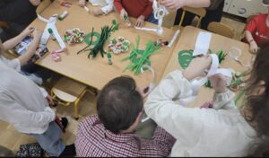 Na zdjęciu widać grupę dzieci i dorosłych siedzących oraz stojących wokół stołu, zajętych pracami plastycznymi. Na stole leżą różne materiały do tworzenia ozdób – zielone paski papieru, kleje w sztyfcie, nożyczki, kolorowe elementy dekoracyjne oraz małe talerzyki z drobnymi przedmiotami, prawdopodobnie słodyczami lub materiałami do ozdabiania. Uczestnicy wydają się skupieni na wycinaniu i sklejaniu papierowych elementów. Atmosfera wygląda na radosną i twórczą, typową dla zajęć plastycznych w szkole lub przedszkolu.