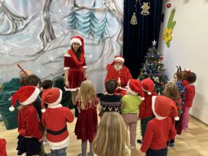 Na zdjęciu widać grupę dzieci uczestniczących w świątecznym spotkaniu z Mikołajem. Dzieci stoją w półkolu, zwrócone w stronę Mikołaja i jego pomocnicy, którzy są ubrani w czerwone stroje świąteczne. Większość dzieci ma na głowach czerwone czapki Mikołaja, a niektóre noszą opaski z rogami renifera.

W tle znajduje się ozdobiona choinka z kolorowymi bombkami i światełkami, a za nią widać zimową dekorację przedstawiającą ośnieżone drzewa. Cała scena odbywa się w jasnej sali, co nadaje zdjęciu ciepły i radosny, świąteczny klimat.
