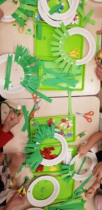 Na zdjęciu widać dzieci wykonujące prace plastyczne przy stole. Na zielonych tackach leżą materiały do tworzenia świątecznych wieńców – papierowe talerzyki z wyciętym środkiem, do których przyklejane są paski zielonego papieru, przypominające gałązki choinki. Obok znajdują się nożyczki, kleje w sztyfcie oraz kolorowe ozdoby, takie jak papierowe gwiazdki i małe dekoracyjne elementy. Dzieci pracują z zaangażowaniem, przyklejając i układając papierowe paski. Cała scena ma radosny, twórczy i świąteczny charakter