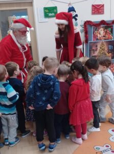 Na zdjęciu widać grupę małych dzieci zgromadzonych wokół Świętego Mikołaja i jego pomocnicy. Mikołaj ubrany jest w tradycyjny czerwony strój z białym futerkiem, czapkę i okulary. Obok niego stoi kobieta w czerwonej sukience z białymi wykończeniami i czapce Mikołaja, pochylona w stronę dzieci z uśmiechem.

Dzieci stoją blisko siebie, zwrócone w stronę Mikołaja, ciekawe tego, co im pokazuje. Ubrane są kolorowo — widać m.in. czerwone, niebieskie i białe ubrania, a niektóre mają świąteczne elementy stroju. W tle znajduje się dekoracja z motywem świątecznym: obrazek z choinką, światełkami i ozdobami, a także napisy i girlandy. Scena odbywa się w jasnej sali przedszkolnej, tworząc ciepłą, radosną i świąteczną atmosferę.