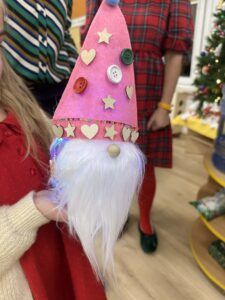 Na zdjęciu widać dziecko trzymające w dłoni świątecznego skrzata wykonanego z materiałów plastycznych. Skrzat ma różowy, stożkowaty kapelusz ozdobiony guzikami, drewnianymi serduszkami i gwiazdkami, a także błyszczącą tasiemką z drobnymi koralikami. Z pod czapki wystaje długa, biała broda z miękkiego, sztucznego włosia, a na środku znajduje się mały, drewniany nosek. W tle widać choinkę oraz osoby ubrane w świąteczne stroje, co nadaje całej scenie ciepły, bożonarodzeniowy klimat