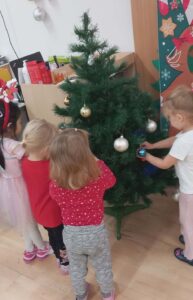Na zdjęciu widać czworo małych dzieci ubierających choinkę. Dzieci stoją blisko drzewka, zawieszając na jego gałązkach kolorowe bombki — głównie złote i niebieskie.
Jedno z dzieci trzyma niebieską bombkę, inne skupione są na ozdabianiu dolnych gałązek. Dzieci ubrane są w jasne i czerwone ubrania, a jedno z nich ma opaskę z rogami renifera. W tle widać świąteczną dekorację na ścianie oraz pudełka i ozdoby. Scena ma ciepły, radosny i świąteczny charakter, typowy dla przygotowań do Bożego Narodzenia w przedszkolu.