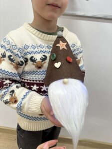 Na zdjęciu widać dziecko trzymające w dłoniach świątecznego skrzata. Skrzat ma brązową, filcową czapkę ozdobioną drewnianą gwiazdką, serduszkiem oraz kolorowymi guzikami w odcieniach czerwieni i zieleni. Na czubku czapki znajduje się mała, szara kulka, a spod niej wystaje długa, biała broda z miękkiego, puszystego materiału. Dziecko ma na sobie kremowy sweter z zimowym wzorem przedstawiającym renifery i śnieżynki w kolorach brązowym, granatowym i bordowym. Cała scena ma ciepły, świąteczny i radosny charakter.