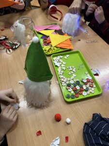 Na zdjęciu widać dzieci wykonujące świąteczne skrzaty podczas zajęć plastycznych. Na stole leży zielona tacka z różnokolorowymi guzikami, drewnianymi gwiazdkami i drobnymi ozdobami. Obok znajdują się kolorowe kartki, nożyczki i inne materiały potrzebne do pracy. Na pierwszym planie stoi gotowy skrzat z długą, białą brodą i zieloną czapką zakończoną białym pomponem. Wokół widać fragmenty filcu, pomponiki i kawałki papieru. Dzieci pracują z zaangażowaniem, tworząc własne dekoracje w świątecznym, radosnym klimacie