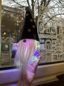 Na zdjęciu widać rękę trzymającą świątecznego skrzata. Skrzat ma długą, białą brodę z miękkiego włosia oraz czarną, stożkowatą czapkę ozdobioną żółtymi gwiazdkami, czerwonym serduszkiem i niebieskim pomponem na czubku. Jego głowa jest przezroczysta i wypełniona kolorowymi światełkami LED, które świecą w odcieniach różu, zieleni, niebieskiego i fioletu, tworząc efektowny, magiczny blask. W tle widać okno z zimową dekoracją przedstawiającą domek, choinkę i śnieżynki, a za szybą – drzewa i fragment ulicy. Całość ma ciepły, świąteczny i nastrojowy klimat.