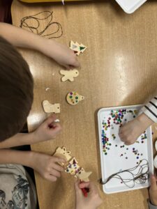 Na zdjęciu widać dzieci dekorujące drewniane ozdoby świąteczne. Na stole leżą zawieszki w kształcie choinki, pierniczka, bombki, misia i skarpety. Dzieci ozdabiają je kolorowymi cekinami, naklejkami i markerami. Na białej tacy obok znajdują się rozsypane błyszczące cekiny w różnych kolorach i kształtach – gwiazdki, kółka i serduszka – oraz kilka sznurków do zawieszenia ozdób. Cała scena przedstawia twórcze, radosne zajęcia plastyczne o świątecznym charakterze