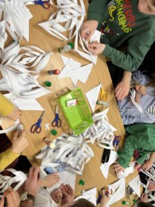 Na zdjęciu widać grupę dzieci siedzących przy stole i wykonujących papierowe ozdoby. Na blacie leży wiele białych pasków papieru, z których powstają przestrzenne śnieżynki lub gwiazdy. Dzieci używają nożyczek, kleju w sztyfcie i zszywacza, aby łączyć poszczególne elementy. Na środku stołu znajduje się zielony pojemnik z przyborami plastycznymi. Uczestnicy są skupieni na pracy, a atmosfera wygląda na radosną i twórczą — typową dla świątecznych zajęć plastycznych.