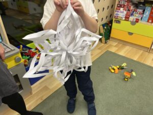 Na zdjęciu widać dziecko trzymające dużą, przestrzenną śnieżynkę wykonaną z białego papieru. Ozdoba składa się z wielu połączonych ze sobą pasków papieru, tworzących efektowny, trójwymiarowy kształt przypominający gwiazdę. Dziecko ma na sobie białą koszulkę, granatowe spodnie i ciemne kapcie. W tle widać fragment sali zabaw lub przedszkola – na podłodze leżą zabawki, a przy ścianie stoją kolorowe szafki i pudełka z grami. Cała scena ma radosny, kreatywny i dziecięcy charakter.