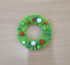 Na zdjęciu znajduje się mały, ręcznie wykonany wieniec świąteczny. Zrobiony jest z zielonych drucików kreatywnych, które zostały owinięte wokół okrągłej podstawy, tworząc puszystą, miękką strukturę. Wieniec ozdobiony jest kolorowymi pomponikami w barwach czerwonej, białej, zielonej i niebieskiej, przypominającymi bombki choinkowe. Całość leży na jasnobrązowym, drewnianym blacie. Dekoracja wygląda prosto, ale bardzo efektownie i ma świąteczny, radosny charakter.