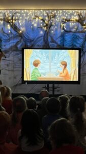 Na zdjęciu widać grupę dzieci siedzących przed dużym ekranem, na którym wyświetlona jest ilustracja przedstawiająca dwoje dzieci siedzących przy stole i pijących coś ciepłego. Za oknem widać zimowy krajobraz – drzewa pokryte śniegiem i błękitne niebo.

Tło sceny, na której odbywa się pokaz, jest ozdobione materiałem z motywem zimowego lasu oraz światełkami przypominającymi gwiazdy lub śnieg. Całość tworzy przytulną, magiczną atmosferę, idealną do zimowego przedstawienia lub zajęć tematycznych w przedszkolu.

Dzieci siedzą spokojnie, zwrócone w stronę ekranu, co sugeruje, że oglądają bajkę, prezentację lub słuchają opowieści o zimie