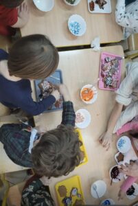 Na zdjęciu widać grupę dzieci siedzących przy stole i dekorujących pierniczki. Na blatach leżą kolorowe tacki z ciastkami w różnych kształtach – gwiazdek, serc czy choinek. Dzieci używają kolorowych lukrów i posypek, które znajdują się w małych miseczkach. Widać pomarańczowy, biały i fioletowy lukier oraz niebieskie i kolorowe ozdoby cukrowe. Atmosfera wygląda na radosną i twórczą – dzieci skupione są na swojej pracy, a stół pełen jest świątecznych kolorów i słodkich dekoracji.