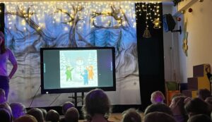 Na zdjęciu widać salę przedszkolną przygotowaną do zimowego przedstawienia lub zajęć tematycznych. Na środku stoi duży ekran, na którym wyświetlony jest obrazek przedstawiający troje dzieci w kolorowych kombinezonach stojących przy bałwanku w ośnieżonym lesie.

Tło sceny ozdobione jest materiałem z namalowanymi drzewami pokrytymi śniegiem, a nad nimi zawieszone są ciepłe, żółte światełka przypominające gwiazdy lub śnieżne iskierki. Po bokach widać elementy dekoracyjne – złotą dzwoneczkową ozdobę oraz kolorowe klocki i meble przedszkolne.

Przed sceną siedzi grupa dzieci, które z zainteresowaniem patrzą w stronę ekranu. Cała scena emanuje przytulną, zimową atmosferą i radością wspólnego oglądania lub uczestniczenia w wydarzeniu.