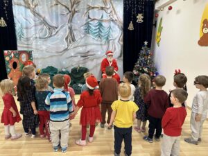 Na zdjęciu widać grupę małych dzieci stojących w półkolu przed osobą przebraną za Świętego Mikołaja. Dzieci są zwrócone do niego plecami, ubrane kolorowo — wiele z nich ma czerwone elementy stroju, a niektóre noszą czapki Mikołaja lub opaski z rogami renifera