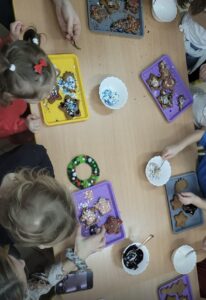 Na zdjęciu widać grupę dzieci siedzących przy stole i dekorujących pierniczki. Na stole leży kilka kolorowych tacek – żółta, fioletowe i szara – na których ułożone są pierniczki w różnych kształtach, m.in. gwiazdek, serc i ludzików. Dzieci ozdabiają je kolorowym lukrem i posypkami, które znajdują się w białych miseczkach. Widać też małą łyżeczkę oraz miseczkę z ciemnym lukrem. Pośrodku stołu leży mały, zielony wieniec z pomponików, przypominający świąteczną ozdobę. Atmosfera wygląda na radosną i twórczą – dzieci skupione są na dekorowaniu ciasteczek, a stół pełen jest kolorów i świątecznego nastroju.