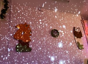 Na zdjęciu widać fragment ściany w przedszkolu lub sali dla dzieci, ozdobionej kolorowymi dekoracjami przedstawiającymi wiewiórkę i sowę. Na środku ściany znajduje się zegar o zielonej ramce.

Całość oświetlona jest efektem świetlnym przypominającym spadające płatki śniegu, które tworzą wrażenie, jakby w pomieszczeniu padał śnieg. Światło ma ciepły, lekko różowy odcień, co nadaje wnętrzu przytulny, zimowy klimat.

W lewym górnym rogu widać fragment zielonej girlandy, co sugeruje, że sala została udekorowana na zimę lub święta. Atmosfera jest radosna i bajkowa, idealna dla dzieci.